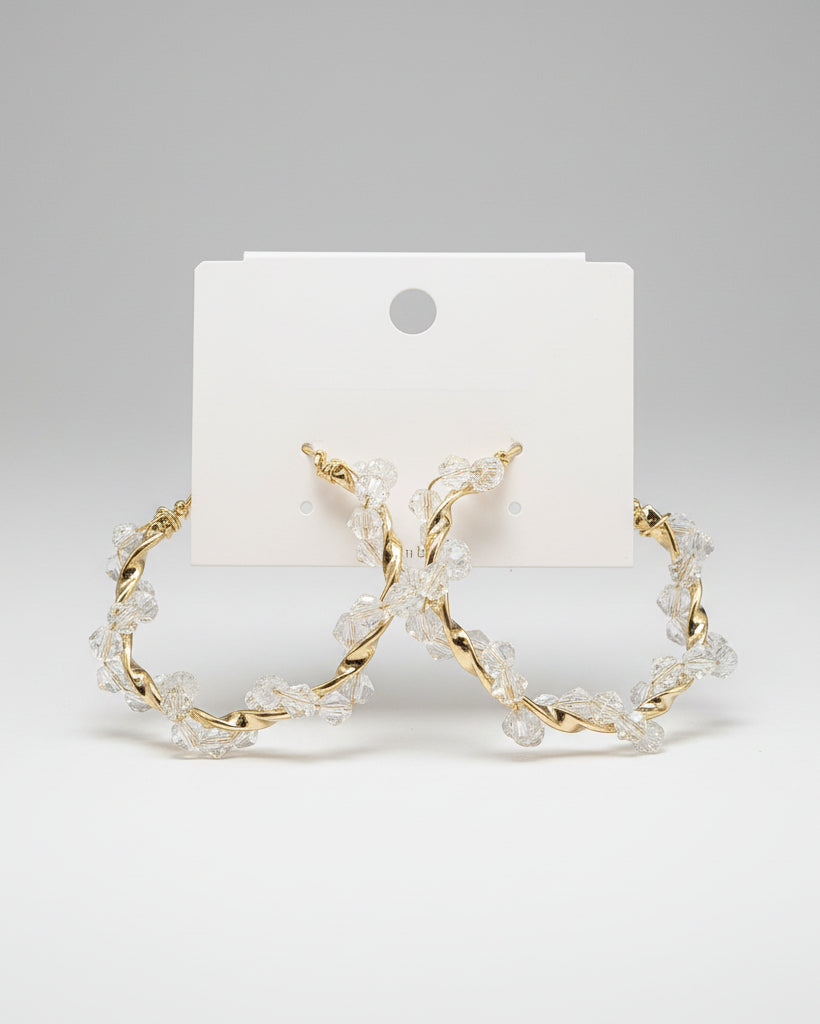 Gold hoop earrings with clear stones on a beige card, placed on a textured fabric background.