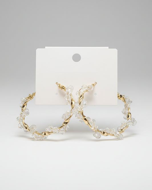 Gold hoop earrings with clear stones on a beige card, placed on a textured fabric background.
