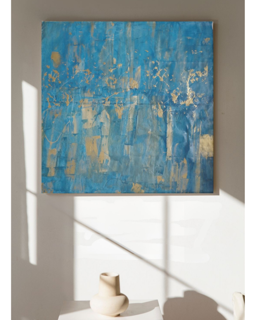 Abstract blue and gold painting on a wall with a white vase below